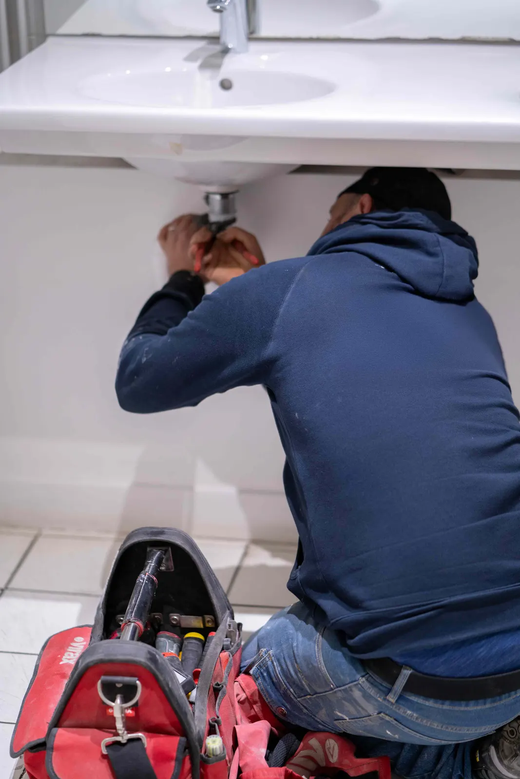 Plombier réparant l'évacuation d'un lavabo, sa sacoche d'outils posée au sol